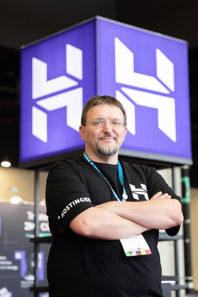 Marco Chiesi in a Hostinger t-shirt, arms folded, in front of a large Hostinger logo