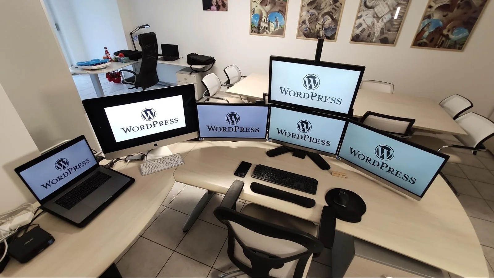 A curved desk with six screens including a laptop, each showing the WordPress wordmark, in a home office with framed pictures on the wall